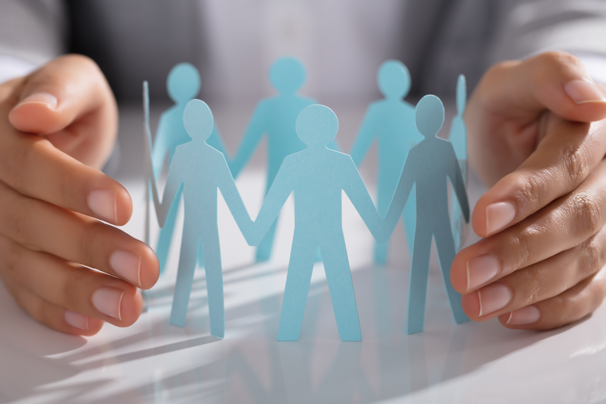 Close-up Of Businessperson Hand Protecting Paper Cut Out Figure On Table In Office