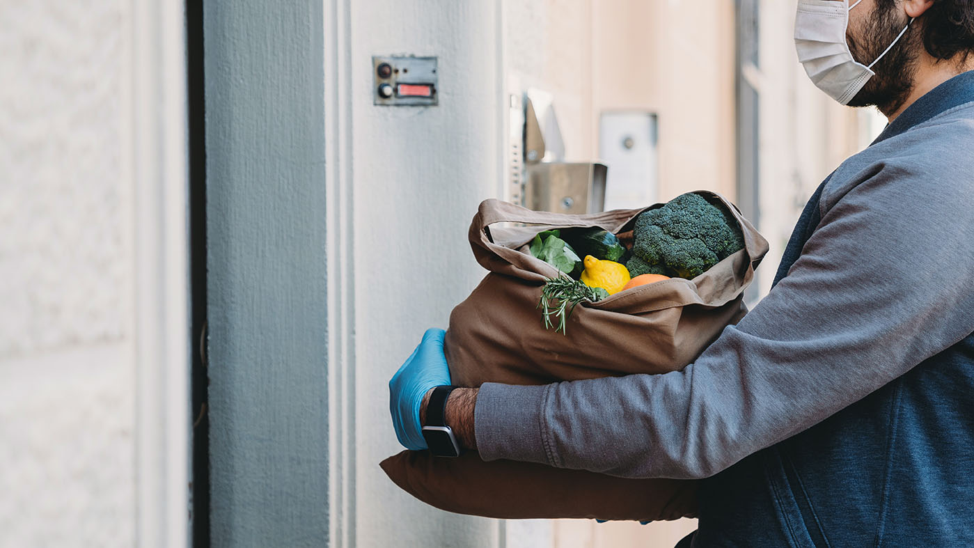 A man is delivering a bag of vegetables and fruit.  He's waiting for the customer to open the door.