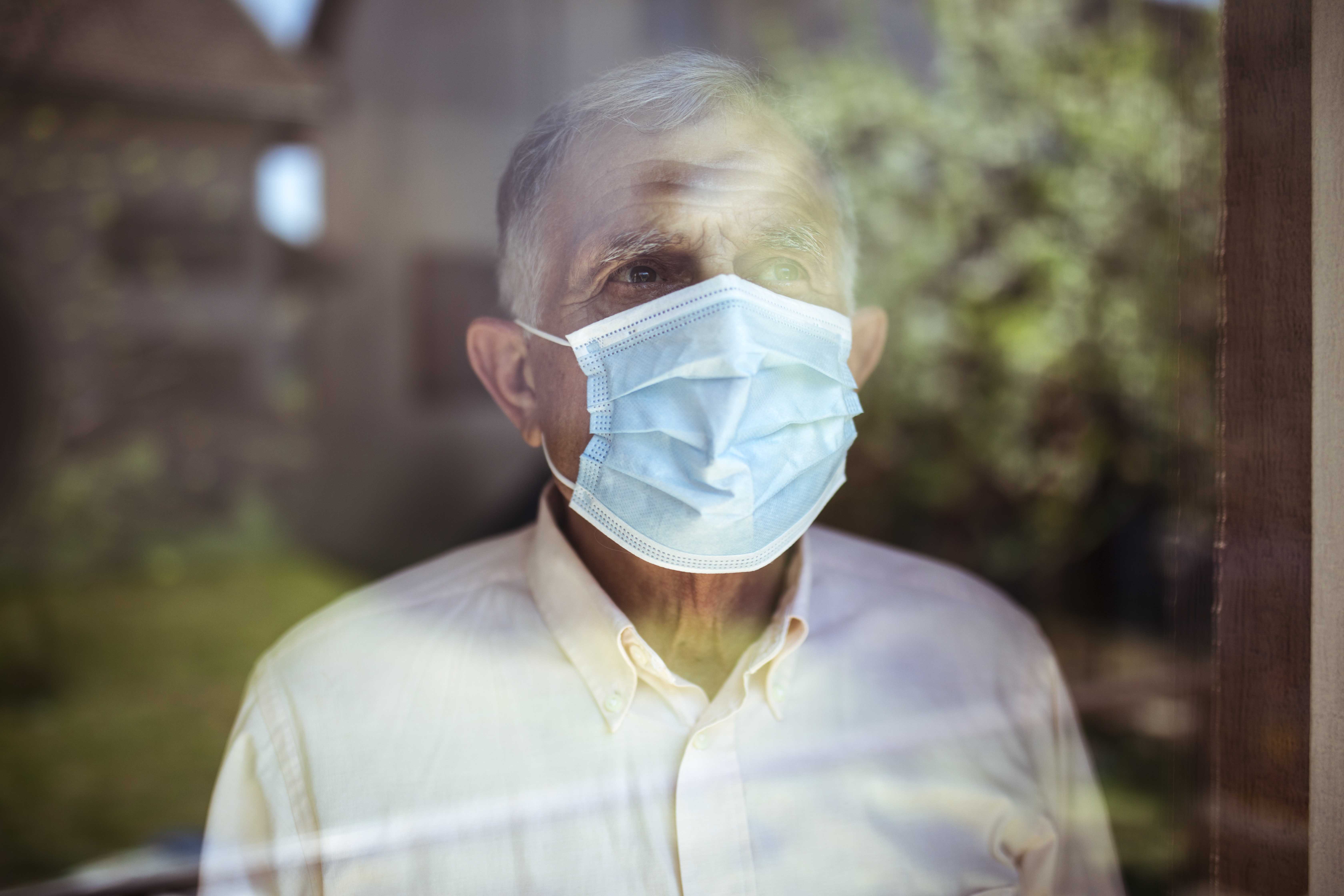 Senior man stays home because of Corona virus. He stands by the window and looks out at the garden.