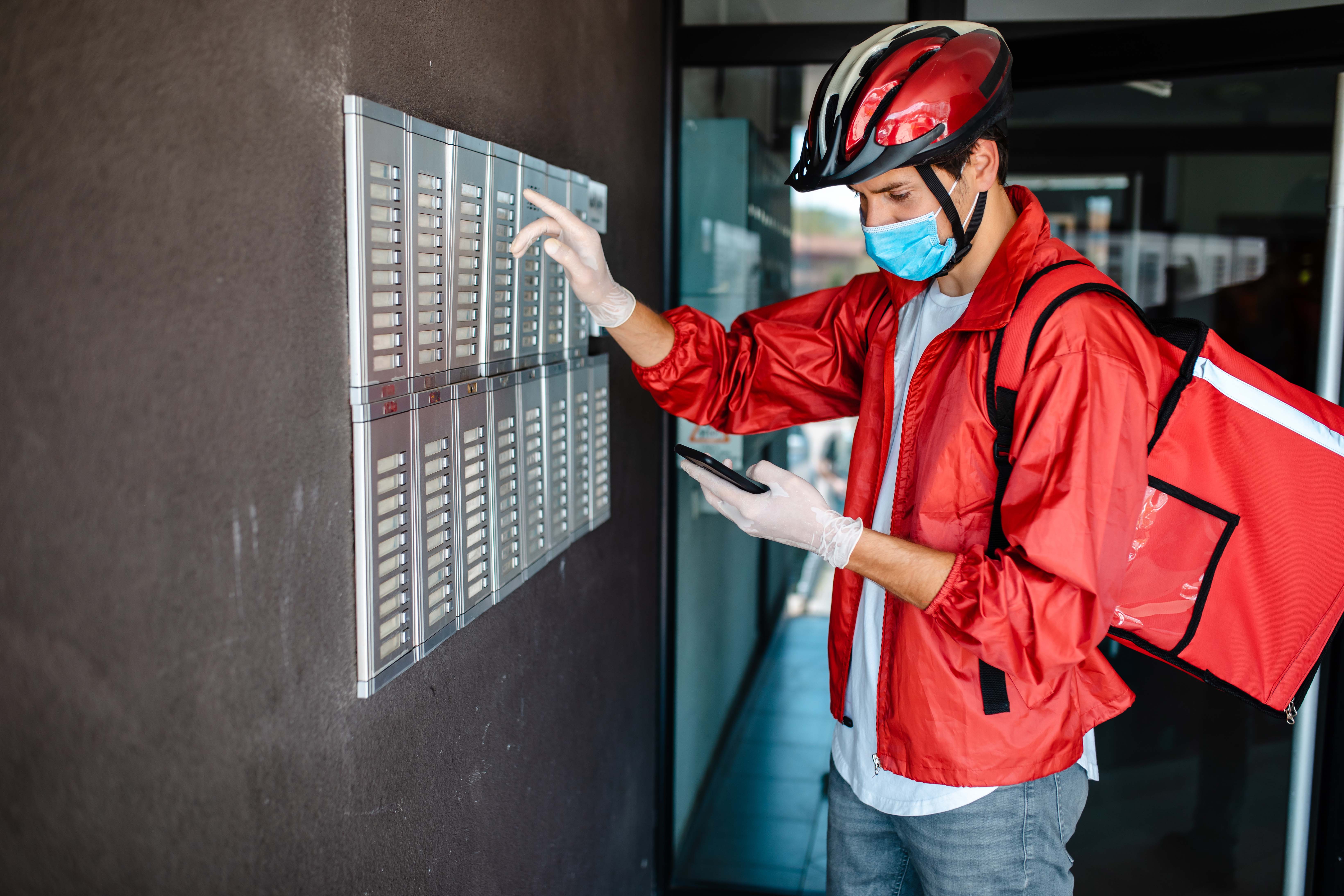 Young delivery person, riniging on intercom while checking the addres on his mobile phone, so he could deliver to a customer on time, while wearing protective gloves and face mask during corona virus pandemic