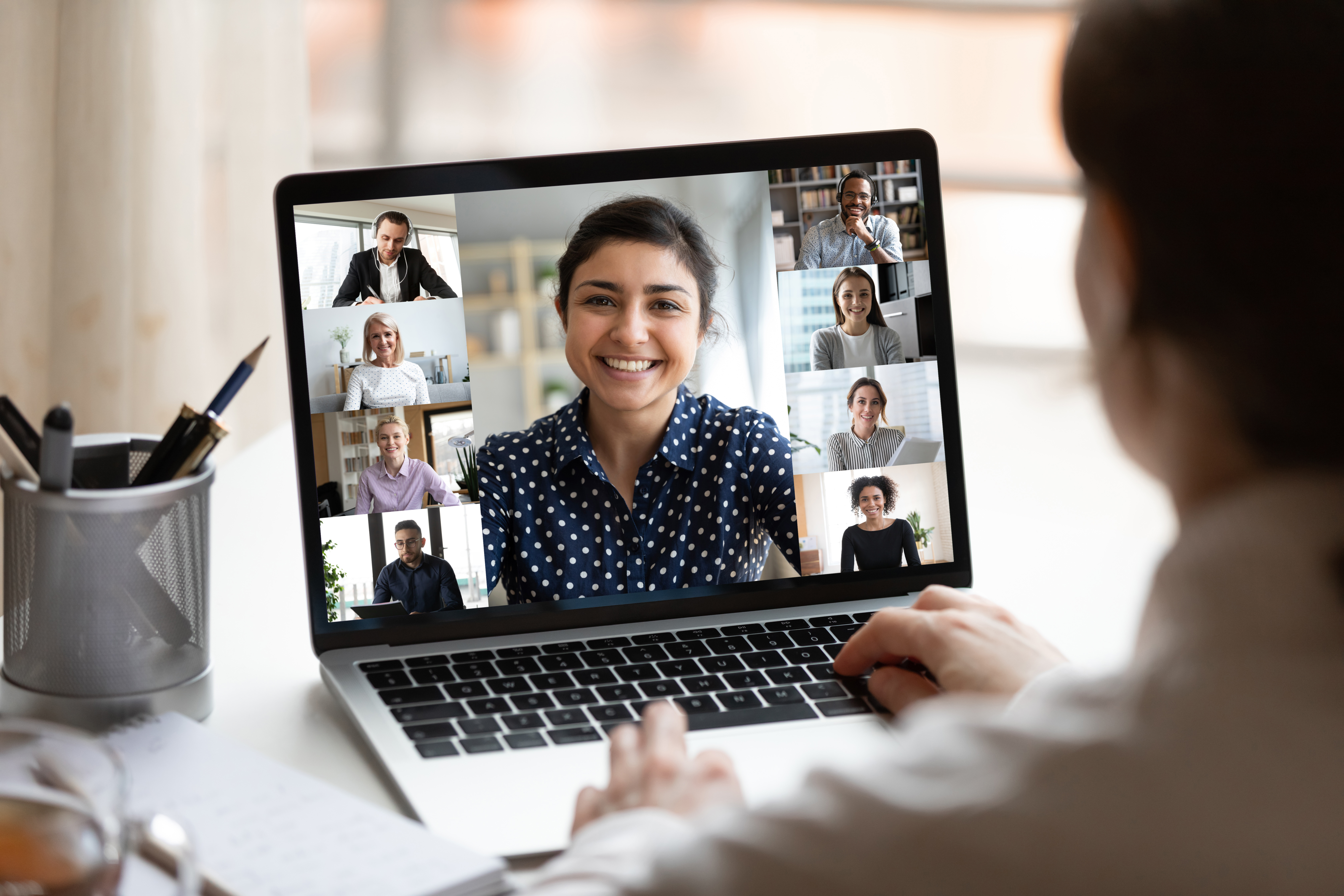 Woman sit at desk looking at computer screen where collage of diverse people webcam view. Indian ethnicity young woman lead video call distant chat, group of different mates using videoconference app