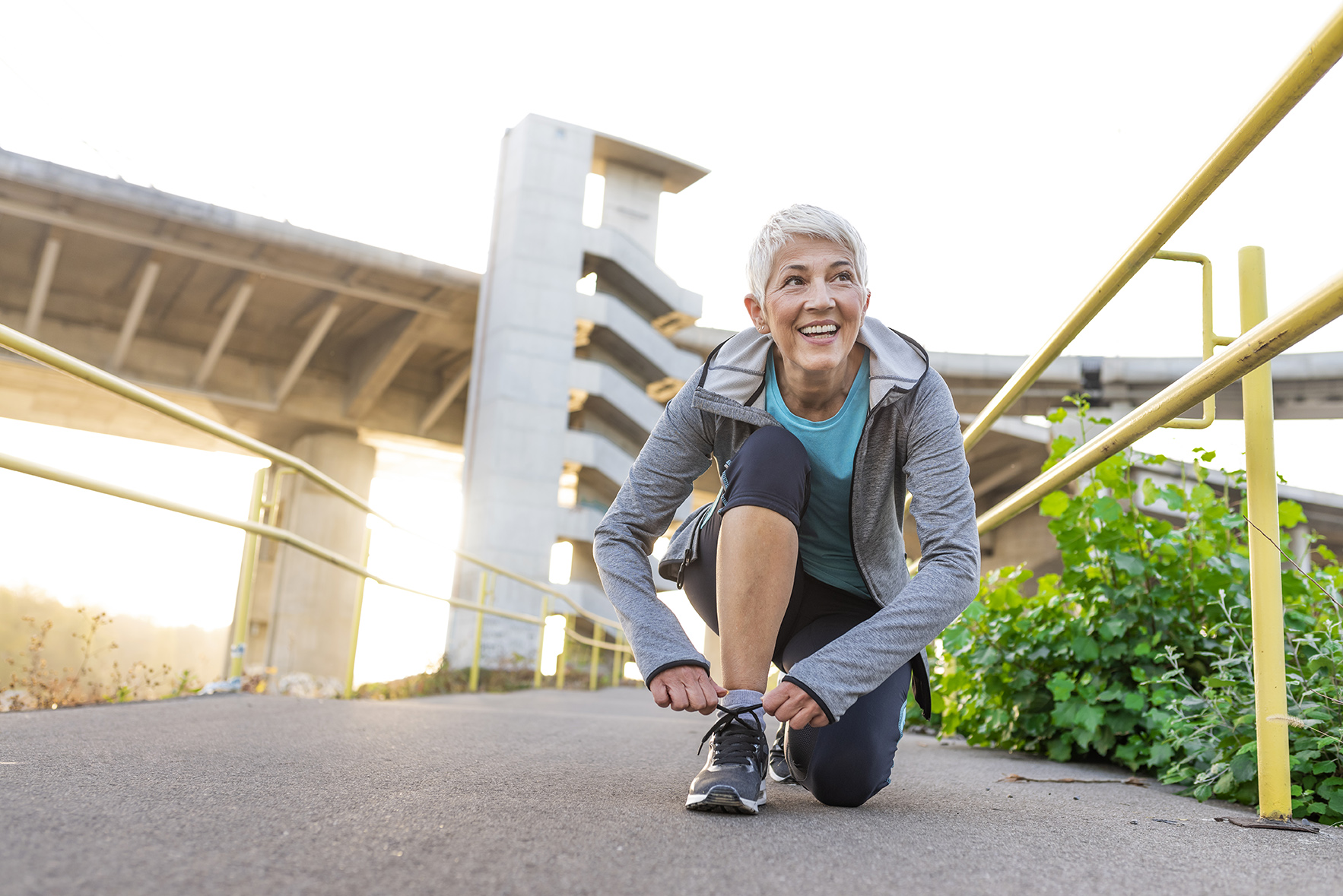 Healthy lifestyle and sport concepts, Photo of Fitness sport mature gray hair woman runner jogging, smiling female runner jogging exercise benefit workout in a Park, tying laces of running shoes before training. Woman tying laces on her sneakers at sunset. Runner having rest while looking away. Sport concept.