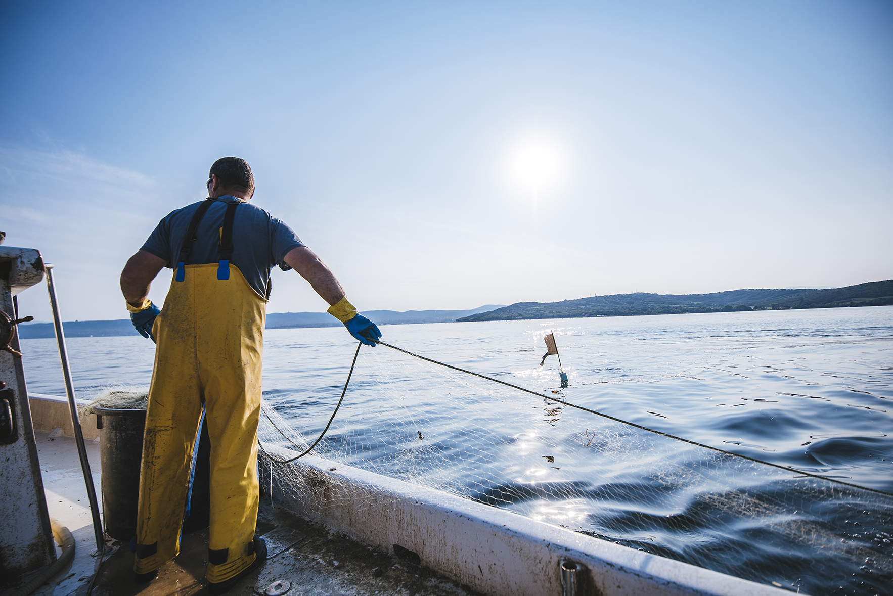 Fisherman putting the fishing net into the water. He is standing on his boat. Sun in back.