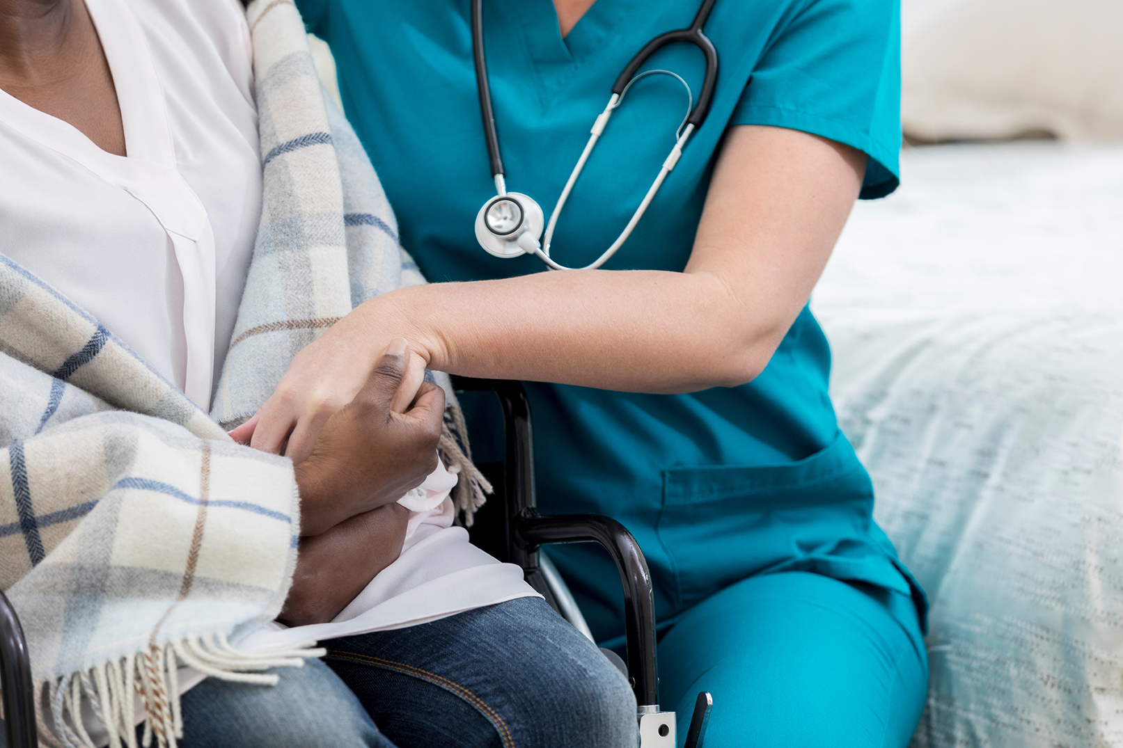 Unrecognizable female home healthcare nurse comforts unrecognizable senior female patient who is wheelchair-bound.