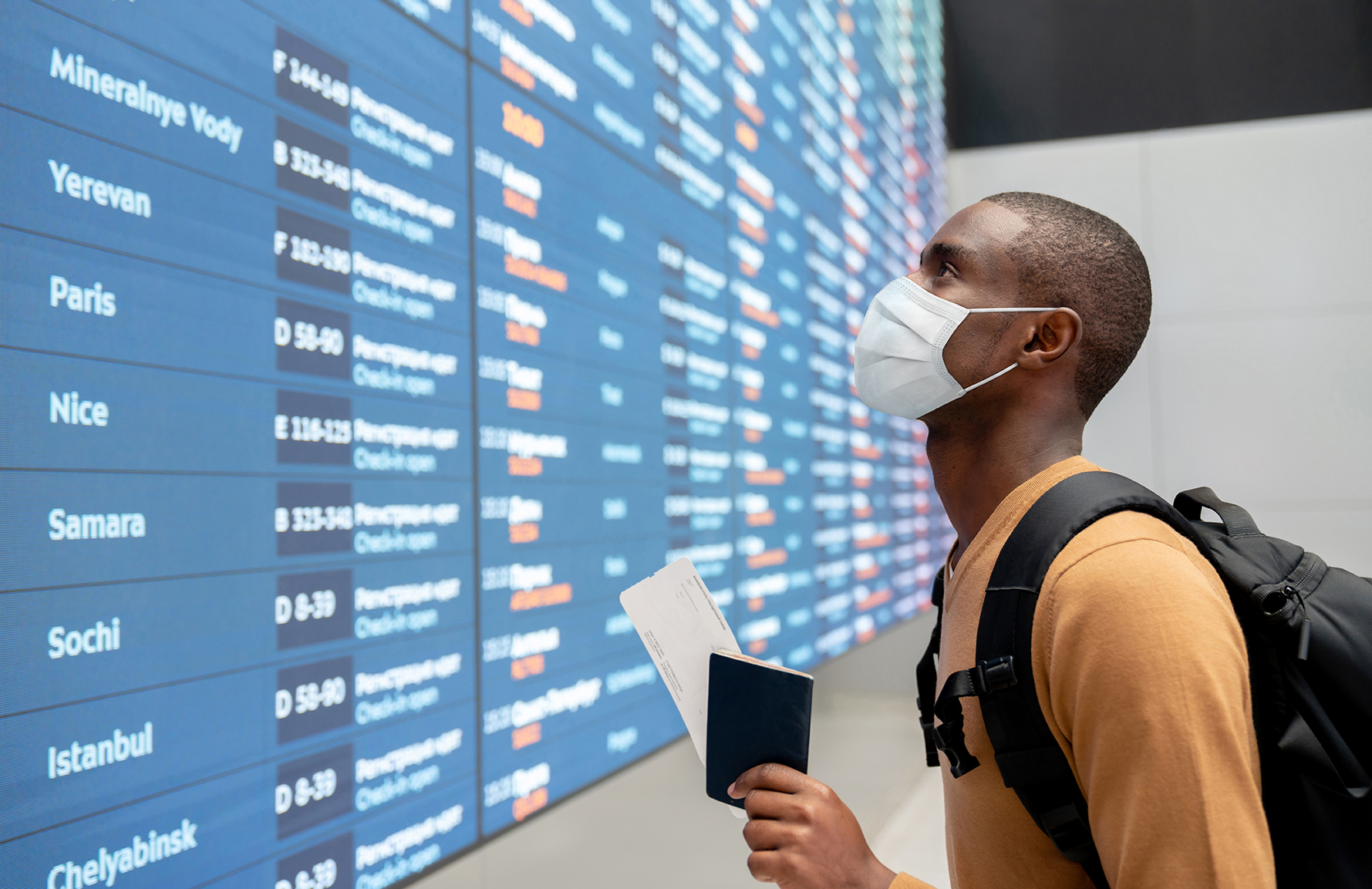 Portrait of a black male traveler wearing a facemask at the airport and looking at the flight schedule - travel concepts