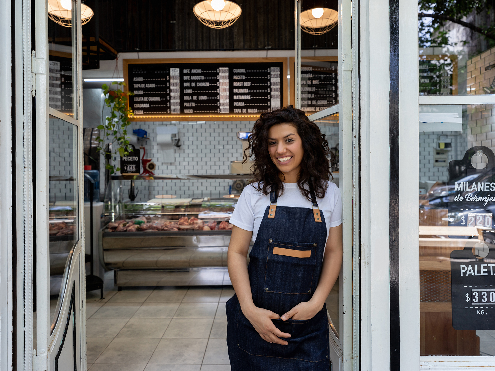 Portrait of beautiful young business owner of a butcher shop leaning against the entrance door smiling at camera very happy