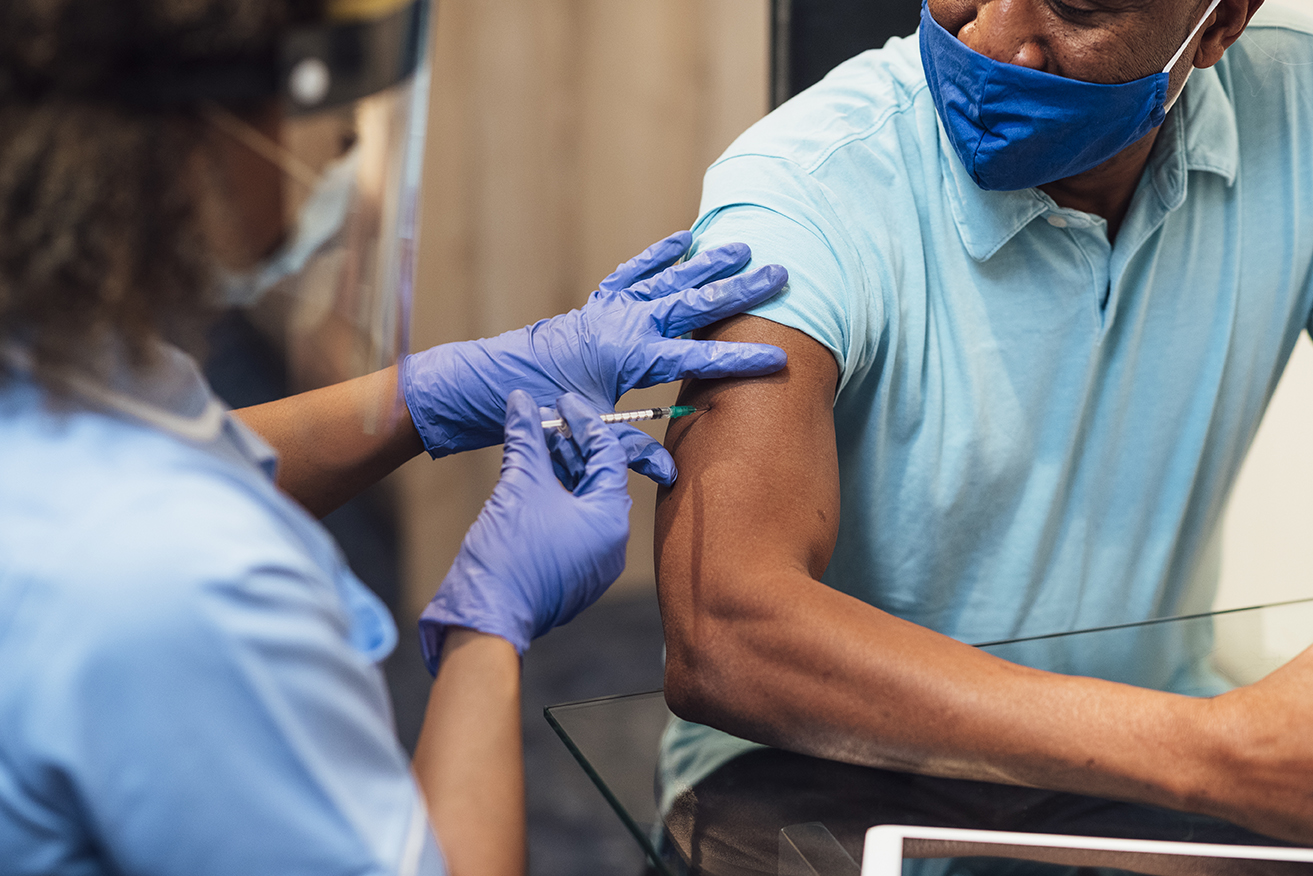 A mixed race female nurse wearing a protective face shield, surgical mask and protective gloves administering the COVID-19 vaccine to a senior black man in his home.