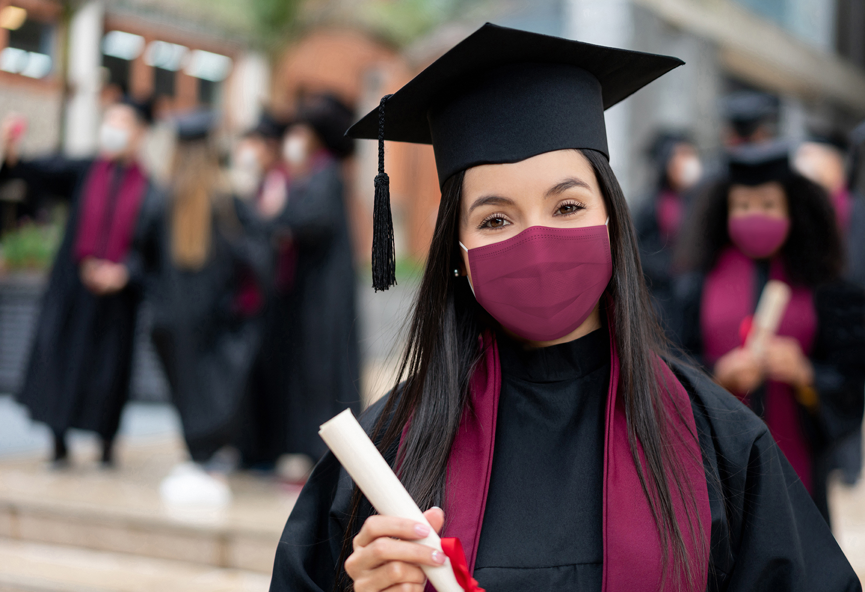 Portrait of a happy graduate student holding her diploma and wearing a facemask at her graduation during the COVID-19 pandemic