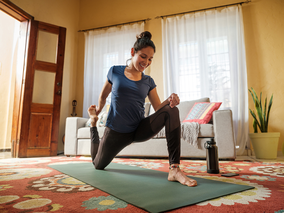 A happy young latin woman stretching her legs on a mat in the living room, holding her leg and smiling.