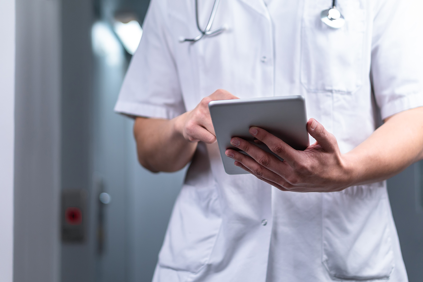 photo of male doctor in uniform with stethoscope coming out of the elevator and using computer tablet in hospital. Modern medical concept