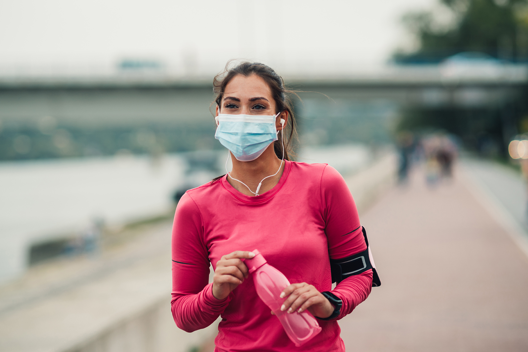 Female runner wearing protective face mask while jogging outdoors during Coronavirus/COVID-19 pandemic. Young woman running in the city next to the river and wearing a protective face mask to protecting from viruses and infections.