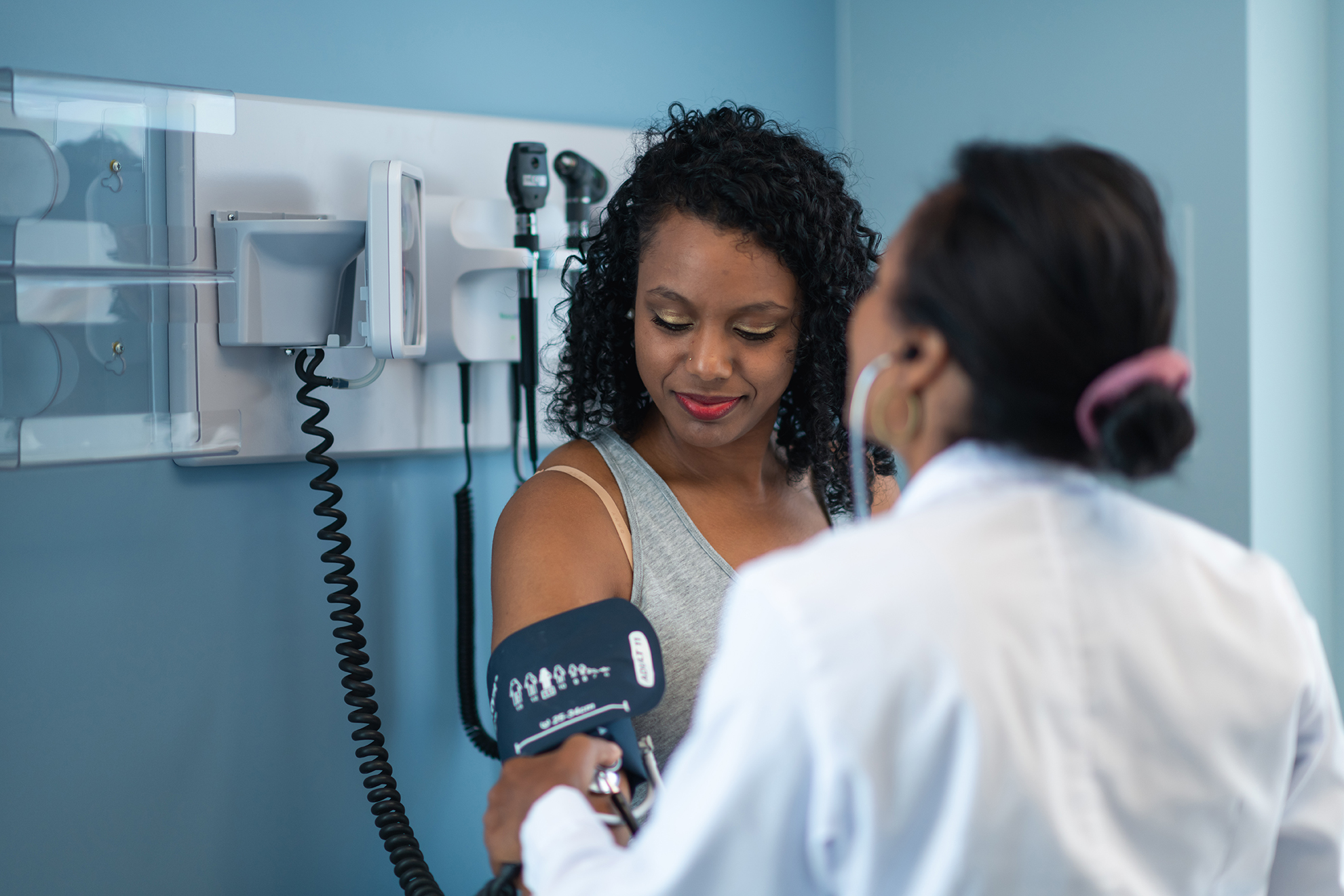 A young black woman is at a routine medical appointment. Her healthcare provider is an ethnic woman. The patient is sitting on an examination table in a clinic. The doctor is checking the patient's blood pressure. The patient is smiling while looking down at her arm.