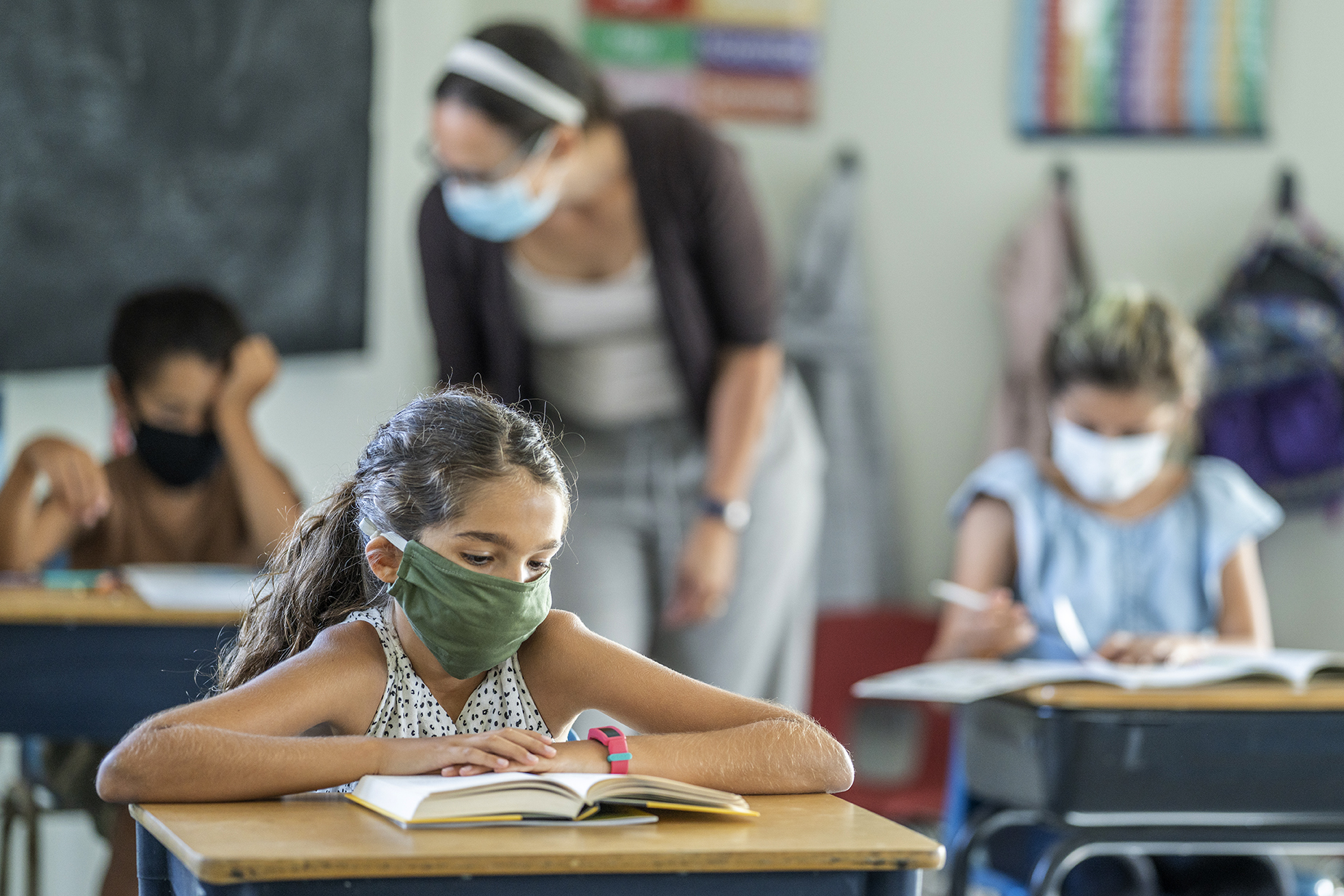12 year old girl wearing a reusable, protective face mask in classroom while working on school work at her desk.