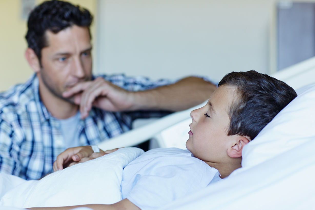Tensed father looking at ill son sleeping in bed at hospital ward
