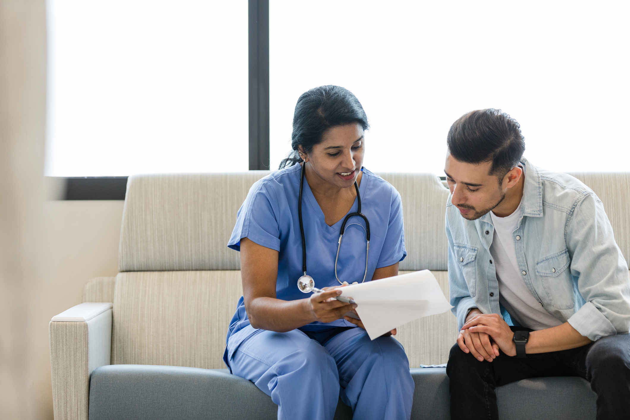 The young man and female doctor sit on the hospital room couch and review the medical chart together.